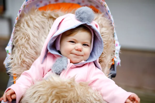 Schattig klein mooi babymeisje zittend in de kinderwagen of kinderwagen op herfstdag. Gelukkig lachend kind in warme kleren, mode stijlvolle roze babyjas met konijnenoren. Baby gaat wandelen met ouders. — Stockfoto