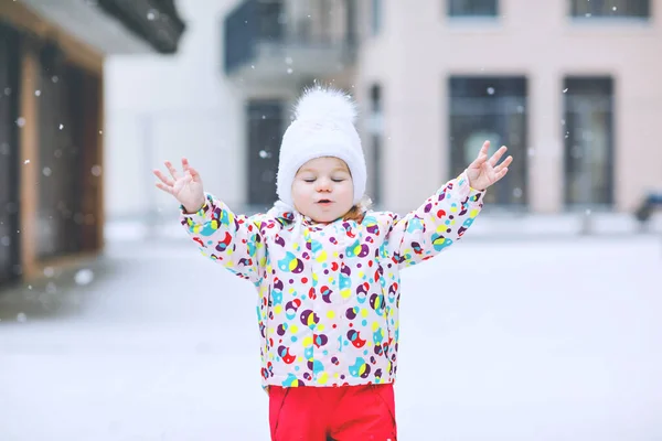 Portret van een klein peutermeisje dat 's winters buiten loopt. Leuke peuter die zoete lolly snoep eet. Kind heeft plezier op koude sneeuwdag. Het dragen van warme baby kleurrijke kleding en hoed met bobbels. — Stockfoto