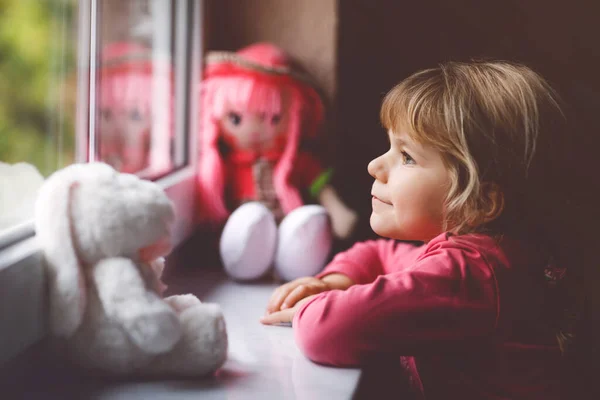 Petite fille mignonne assise à côté de la fenêtre et regardant dehors le jour de pluie. Enfant rêveur avec poupée et jouet doux se sentant heureux. Self isolation concept during corona virus pandemic time. Enfant solitaire. — Photo