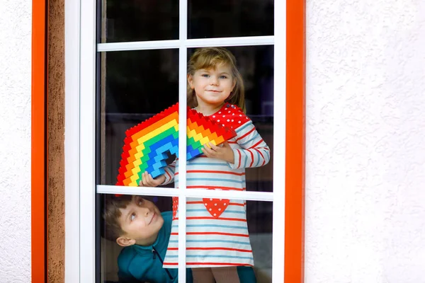Batole dívka a chlapec po okně vytvořit duhu s barevnými plastovými bloky během pandemické koronavirus karantény. Dvě děti, sourozenci a malování duhy po celém světě jako znamení. — Stock fotografie