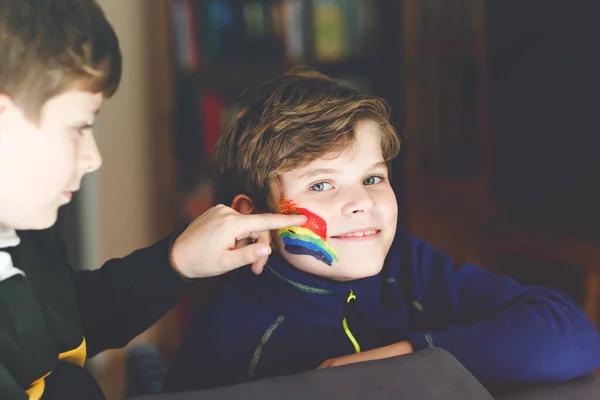 Twee jongens met regenboog met kleurrijke kleuren op het gezicht tijdens pandemische coronavirus quarantaine. kind schildert het gezicht van broer. Kinderen maken regenbogen over de hele wereld. Broers en zussen, beste vrienden — Stockfoto