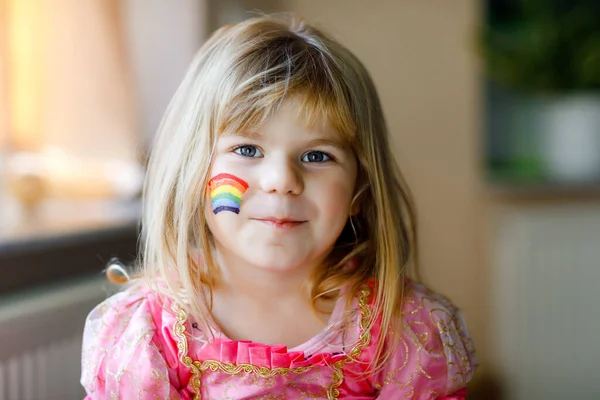 Retrato de linda niña pequeña con arco iris pintado con colores coloridos en la cara. Niño solitario durante la cuarentena del coronavirus pandémico. Los niños hacen y pintan arcoíris en todo el mundo — Foto de Stock