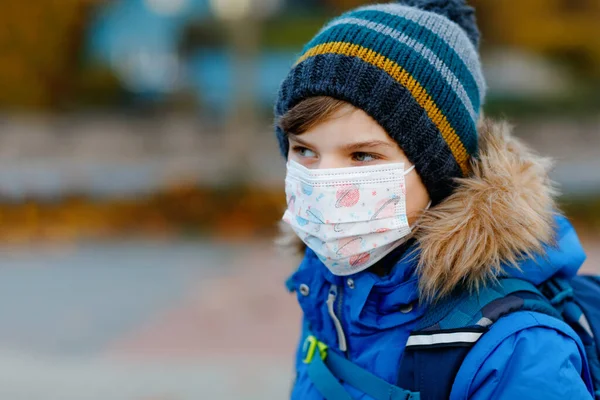 Niño pequeño con máscara médica camino a la escuela. Mochila para niños. Colegial en otoño frío o día de invierno con ropa de abrigo. Tiempo de bloqueo y cuarentena durante la enfermedad pandémica de corona — Foto de Stock