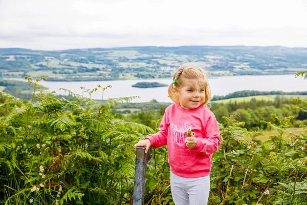 Söt småbarn flicka med irländskt landskap och gröna kullar och sjö på bakgrunden. Glada friska barn njuta av naturen. Familje- och småbarnssemester i Irland — Stockfoto