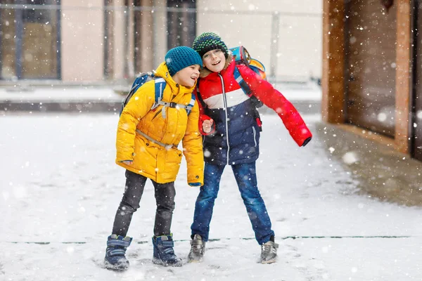 Dwójka małych chłopców z podstawówki, idących do szkoły podczas opadów śniegu. Szczęśliwe dzieci bawiące się i bawiące z pierwszym śniegiem. Rodzeństwo i najlepsi przyjaciele z plecakiem w kolorowe ubrania zimowe. — Zdjęcie stockowe