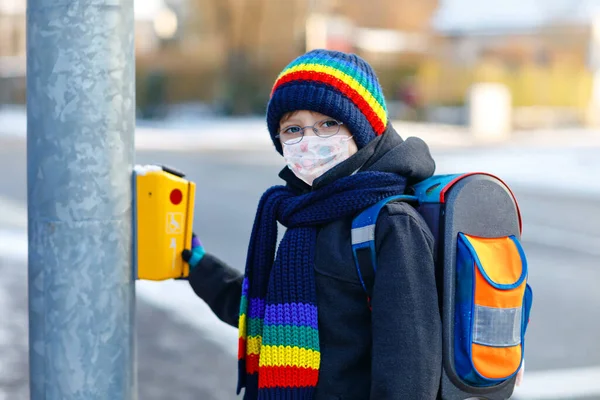 Pieni silmälasipäinen poika lääketieteellisessä naamiossa matkalla kouluun. Lapsi reppu laukku. Koulupoika talvipäivänä lämpimillä vaatteilla. Lukitus ja karanteeniaika koronapandemian aikana — kuvapankkivalokuva