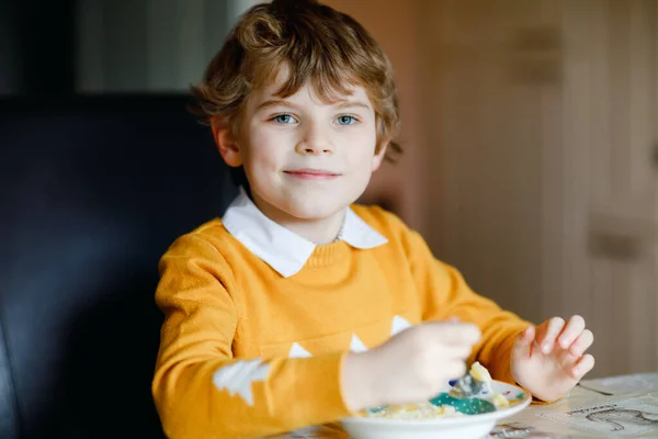 Menino da escola a comer sopa de legumes no interior. Criança loira na cozinha doméstica ou na cantina da escola. Criança bonito e comida saudável, sopa vegan orgânica com macarrão, frango e legumes. — Fotografia de Stock