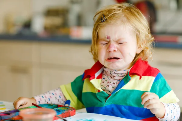 Szomorú síró kislány, aki vízfestékkel tanul festeni. Kisgyermek rajz otthon, színes ecseteket használva. Dühös gyermek hisztérikus válságban. — Stock Fotó