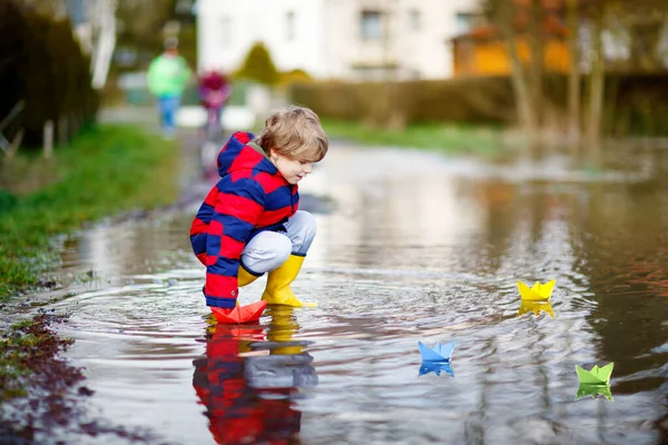 Felice bambino in stivali da pioggia gialli giocare con la barca nave di carta da enorme pozzanghera in primavera o autunno giorno — Foto Stock