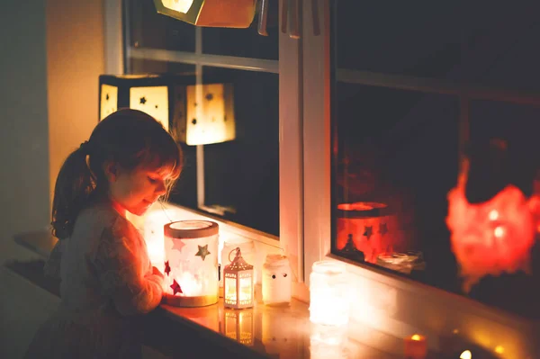 Niña sentada junto a la ventana con linternas hechas a mano con velas para la procesión de San Martín. Un niño pequeño mirando una linterna brillante. Tradición alemana Martinsumzug. Decoración del hogar —  Fotos de Stock