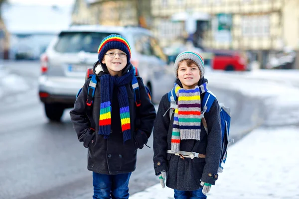 Två små pojkar i grundskolan som går till skolan under snöfallet. Glada barn som har roligt och leker med första snön. Syskon an vän med ryggsäck i färgglada vinterkläder. — Stockfoto