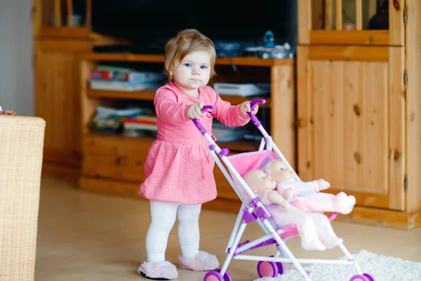 Мила чарівна дівчинка робить перші кроки і штовхає з ляльковою каретою. Красива дитина-малюк штовхає коляску з іграшкою вдома. Щаслива дитина з двома ляльками — стокове фото