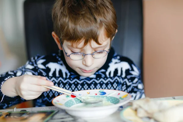 Kleine schooljongen met een bril die binnen groentesoep eet. Blond kind in de huiselijke keuken of in schoolkantine. Schattig kind en gezond eten, biologische veganistische soep met noedels, kip en groenten. — Stockfoto