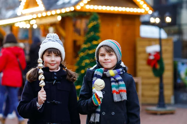 Malá roztomilá holčička a chlapec jíst bílou čokoládu pokryté jahody a jablko na špejli na tradičním německém vánočním trhu. Happy children, best friends, twins and siblings on snowy day — Stock fotografie