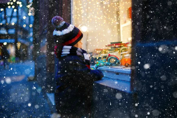 Cute toddler boy looking on car toys in a window on Christmas time season. Fascinated child in winter clothes dreaming and wishing. Window decorated with xmas gifts. Snow falling down, snowfall. — Stock Photo, Image