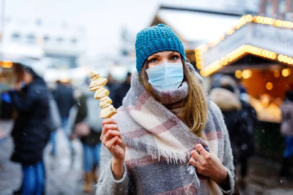 Mujer con máscara médica comiendo frutas cubiertas de chocolate blanco en pincho en el mercado tradicional de Navidad. Personas con máscaras como protección contra el virus corona. Tiempo de pandemia covid. — Foto de Stock