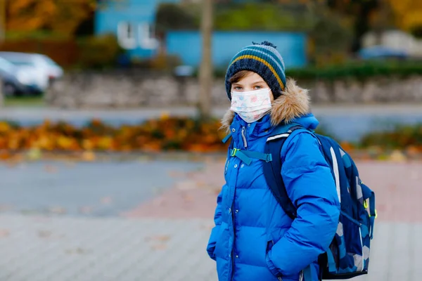 Niño pequeño con máscara médica camino a la escuela. Mochila para niños. Colegial en otoño frío o día de invierno con ropa de abrigo. Tiempo de bloqueo y cuarentena durante la enfermedad pandémica de corona — Foto de Stock