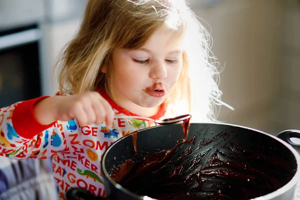 可爱的幼儿小女孩吃巧克力面团，用勺子和手指从锅中休息。快乐的孩子舔甜面团做松饼或蛋糕，在家里厨房帮忙，在室内帮忙. — 图库照片