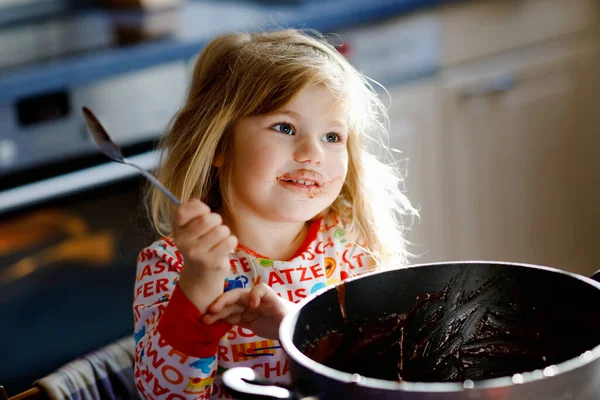 Cute toddler girl eating chocolate dough rests with spoon and fingers from pot. Happy child licking sweet dough for muffins or cake, helping in home kitchen, indoors. — Stock Photo, Image