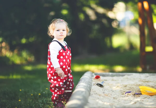 Linda niña jugando en la arena en el patio al aire libre. Hermoso bebé en pantalones de goma roja que se divierten en el soleado día cálido de verano. Niño con juguetes de arena de colores. Bebé activo saludable al aire libre juega juegos —  Fotos de Stock