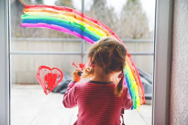 Adoralbe kleine peuter meisje met regenboog en hart geschilderd met kleurrijke venster kleur tijdens pandemische coronavirus quarantaine. Kind schilderen regenbogen over de hele wereld met de woorden Laten we allemaal goed zijn — Stockfoto