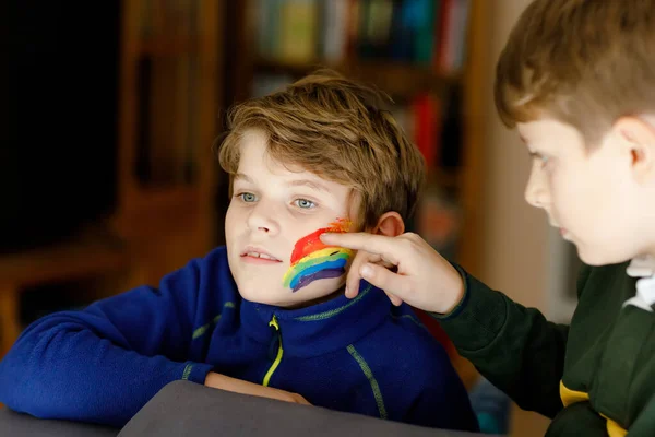 Zwei kleine Jungen mit Regenbogen mit bunten Farben im Gesicht während der Coronavirus-Quarantäne. Kind malen das Gesicht des Bruders. Kinder basteln Regenbogen auf der ganzen Welt. Geschwister, beste Freunde — Stockfoto