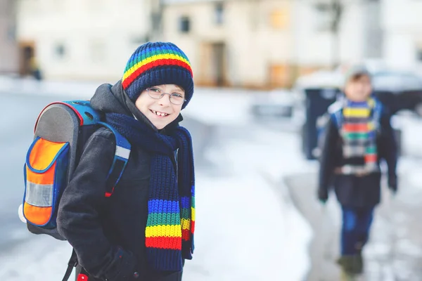 İlköğretim sınıfından iki çocuk kar yağışı sırasında okula yürüyorlar. Mutlu çocuklar eğleniyor ve ilk karla oynuyorlar. Kardeşler, renkli kış kıyafetleri içinde sırt çantalı arkadaşlar.. — Stok fotoğraf