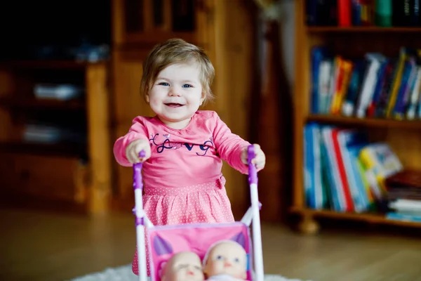 Schattig schattig baby meisje maken eerste stappen en duwen met pop rijtuig. Mooi peuter kind duwen kinderwagen met speelgoed thuis. Gelukkig kind met twee poppen — Stockfoto