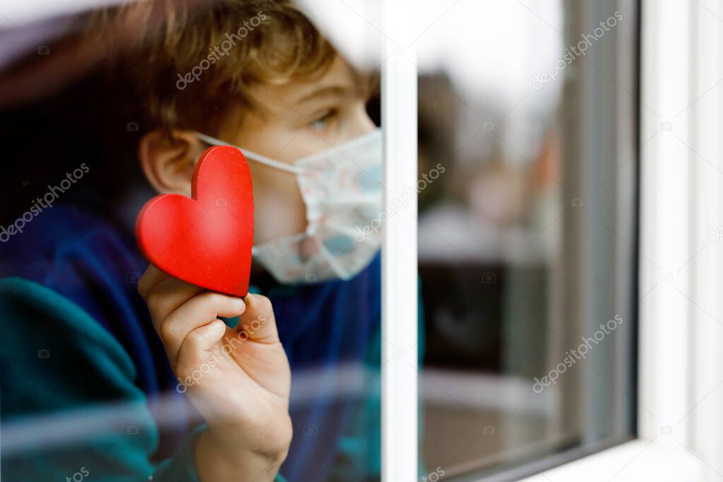 Lovely little school kid boy by a window wearing medical mask and holding wooden heart during pandemic coronavirus quarantine. Lonely upset child, self isolation concept.
