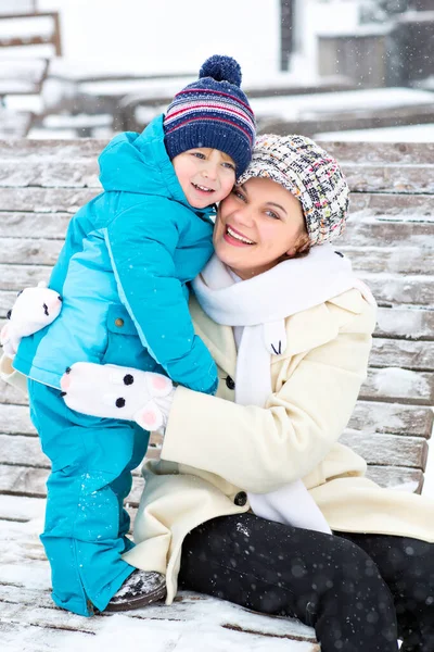 Young mother hugging with toddler boy sitting on bench in winter park. Woman playing and hugging with cute baby son. Adorable kid boy, child in colorful clothes. Snow falls. Family time. — Stock Photo, Image