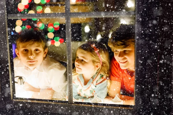 Tre söta barn sitter vid fönstret på julafton. Två skolpojkar och småbarn flicka, syskon ser utomhus och drömmer. Familjens lycka på traditionell semester — Stockfoto