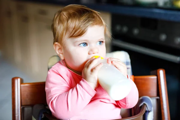 Linda menina adorável segurando mamadeira e bebendo leite fórmula. Primeira comida para bebés. Criança recém-nascida, sentada na cadeira da cozinha doméstica. Bebês saudáveis e conceito de alimentação por mamadeira — Fotografia de Stock