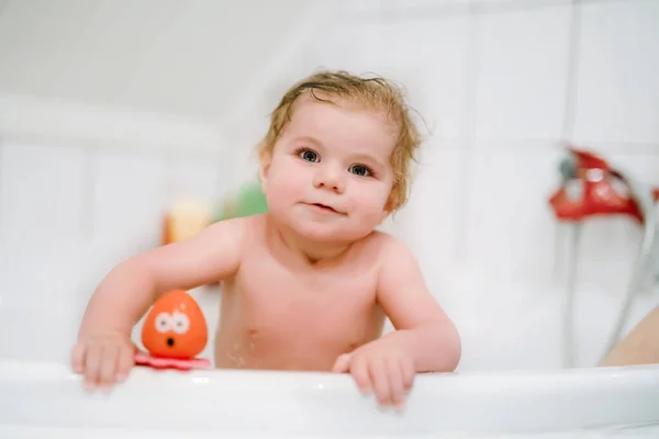 Cute adorable baby girl taking foamy bath in bathtub. Toddler playing with bath rubber toys. Beautiful child having fun with colorful gum toys and foam bubbles — Stock Photo, Image