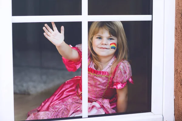 전 세계적 인 코로나 바이러스 격리가 진행되는 동안 공주 옷을 입은 외로워 보이는 작은 아기가 무지개와 함께 창가에 앉아 있다. 아이들은 전 세계에서 무지개를 만들고 페인트칠을 한다 — 스톡 사진