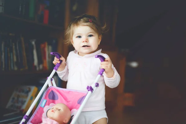 Linda niña adorable haciendo los primeros pasos con carruaje de muñeca. Hermoso niño pequeño empujando cochecito con juguete en casa. Feliz hija aprendiendo a caminar y de pie —  Fotos de Stock