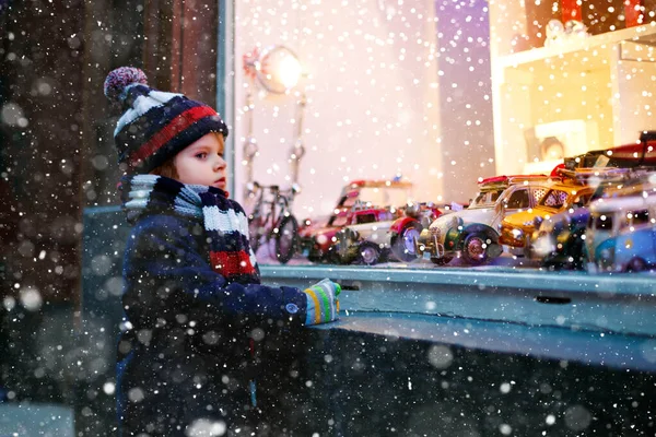 Lindo niño mirando juguetes de coche en una ventana en la temporada navideña. Niño fascinado en ropa de invierno soñando y deseando. Ventana decorada con regalos de Navidad. Nieve cayendo, nieve cayendo. — Foto de Stock