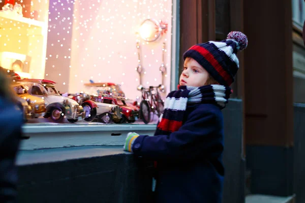 Cute toddler boy looking on car toys in a window on Christmas time season. Fascinated child in winter clothes dreaming and wishing. Window decorated with xmas gifts. Snow falling down, snowfall. — Stock Photo, Image