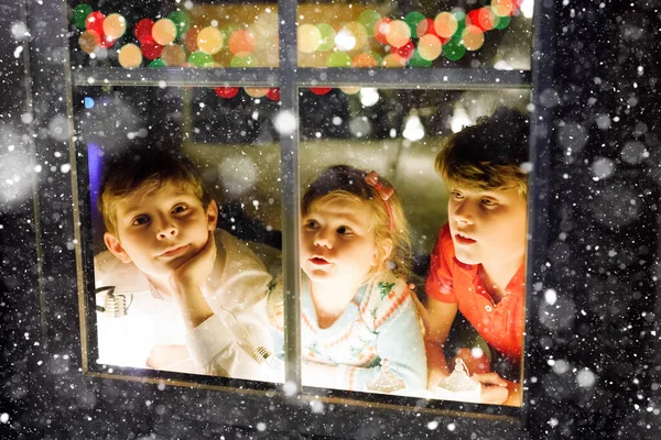 Tři roztomilé děti sedí u okna na Štědrý večer. Dva školáci a batolata, sourozenci, co vypadají venku a sní. Rodinné štěstí na tradiční dovolené — Stock fotografie