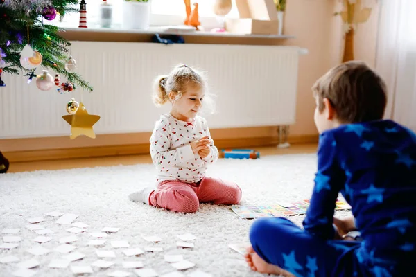 Two little chilren, cute toddler girl and school kid boy playing together card game by decorated Christmas tree. Happy healthy siblings, brother and sister having fun together. Family celebrating xmas — Stock Photo, Image