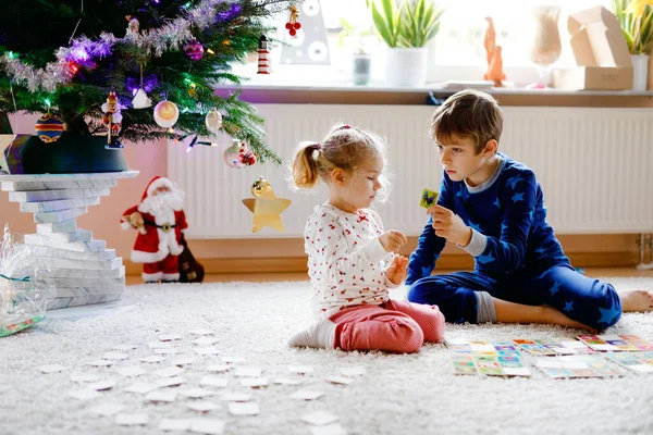 Two little chilren, cute toddler girl and school kid boy playing together card game by decorated Christmas tree. Happy healthy siblings, brother and sister having fun together. Family celebrating xmas — Stock Photo, Image