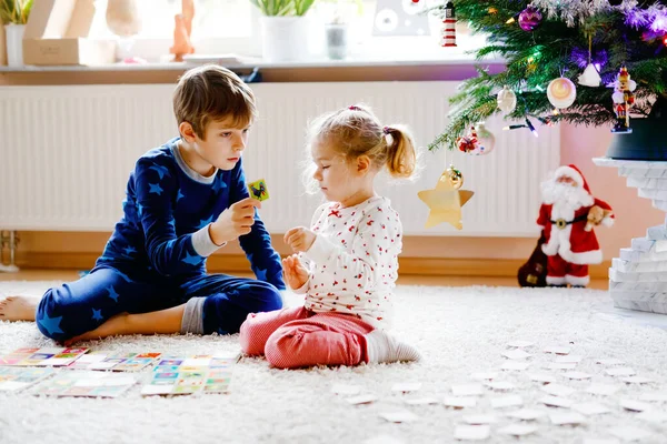 Two little chilren, cute toddler girl and school kid boy playing together card game by decorated Christmas tree. Happy healthy siblings, brother and sister having fun together. Family celebrating xmas — Stock Photo, Image