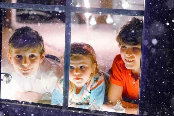Trójka uroczych dzieci siedzących przy oknie w Wigilię. Dwóch chłopców ze szkoły i mała dziewczynka, rodzeństwo wyglądające na zewnątrz i marzące. Szczęście rodzinne na tradycyjnym urlopie — Zdjęcie stockowe