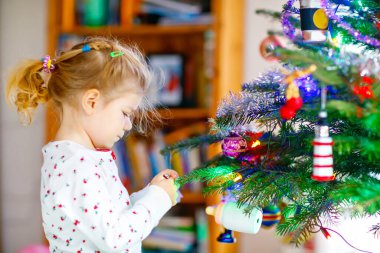Pijamalı sevimli bir kız Noel ağacını şirin ellerle süslüyor. Gecelikli küçük çocuk Noel ağacının yanında duruyor. Geleneksel aile kış tatilinin kutlaması