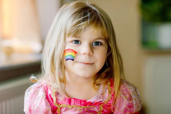 Retrato de linda niña pequeña con arco iris pintado con colores coloridos en la cara. Niño solitario durante la cuarentena del coronavirus pandémico. Los niños hacen y pintan arcoíris en todo el mundo — Foto de Stock