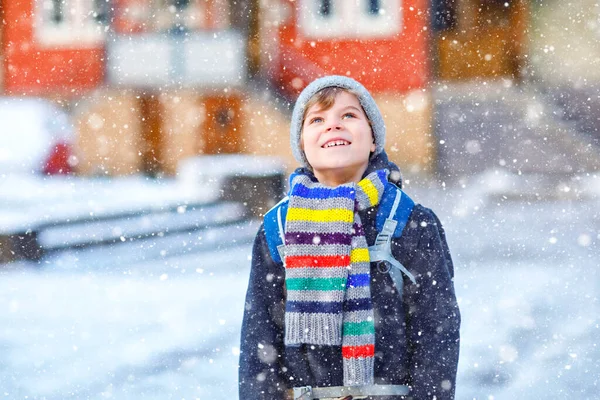 Malý školáček základní třídy, který chodí do školy během sněžení. Šťastné dítě baví a hrát si s prvním sněhem. Student s batohem nebo brašnou v barevných zimních šatech. — Stock fotografie