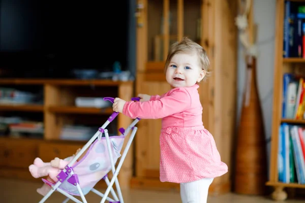 Cute adorable baby girl make first steps and push with doll carriage. Beautiful toddler child pushing stroller with toy at home. Happy kid with two dolls — Stock Photo, Image