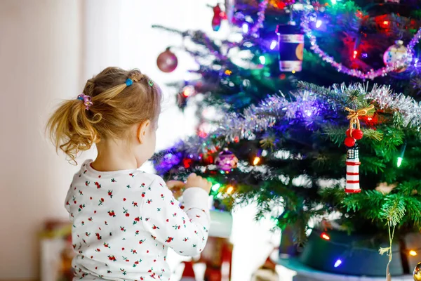 Roztomilé batole dívka v pyžamu zdobí vánoční stromek s hračkou v roztomilých rukou. Malé dítě v noční košili stojící u vánočního stromku. oslava tradiční rodinné zimní dovolené — Stock fotografie