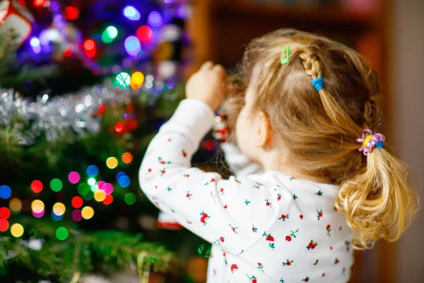 사랑 스러운 잠옷을 입은 아기가 귀여운 손으로 크리스마스 트리를 장식하고 있다. 크리스마스 트리 옆에 서 있는 촌뜨기 꼬마. 전통적 인 가족의 휴일 기념행사 — 스톡 사진