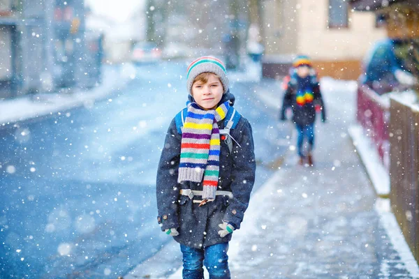 Deux petits garçons de classe primaire marchant jusqu'à l'école pendant les chutes de neige. Enfants heureux s'amuser et jouer avec la première neige. Frères et sœurs et amis avec sac à dos en vêtements d'hiver colorés. — Photo