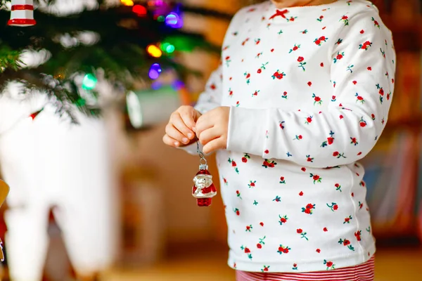 Adorable toddler girl in pajamas decorating Christmas tree with toy in cute hands. Little child in nightwear standing by Xmas tree. celebration of traditional family winter holiday — Stock Photo, Image